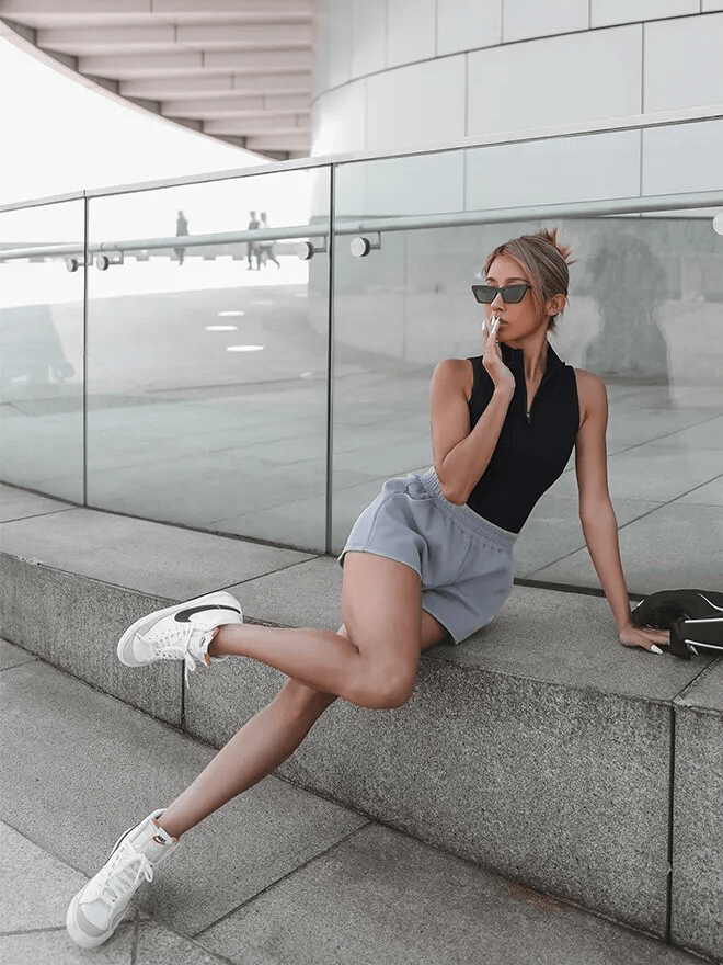 Woman in stylish casual wear sitting on modern stone bench, wearing sunglasses and sneakers, exuding confidence and chic style.